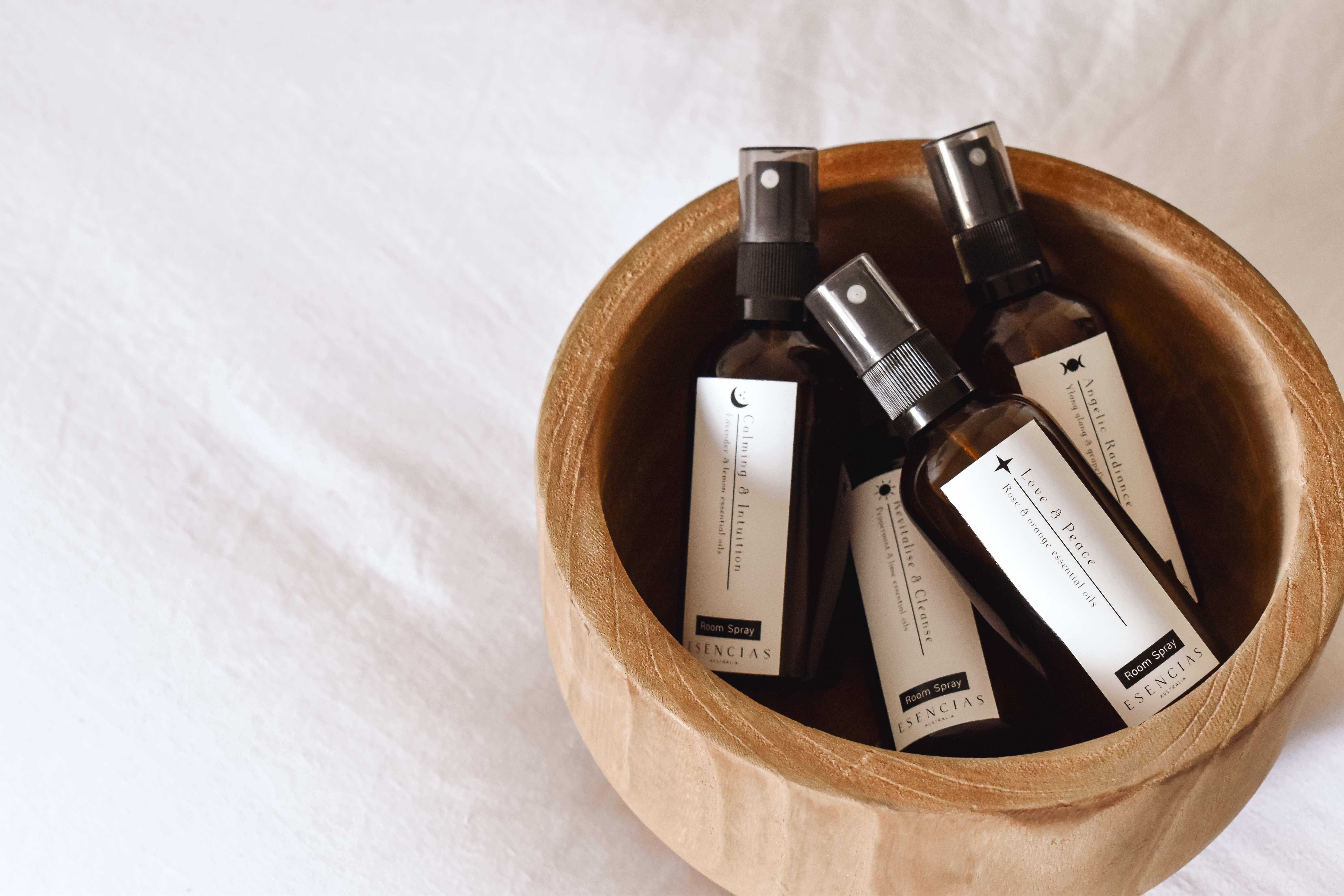 All four Esencias room sprays – Calming & Intuition, Love & Peace, Angelic Radiance and Revitalise & Cleanse – resting in a wooden bowl on soft white linen